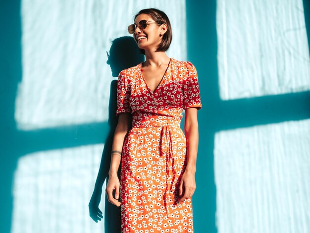 Young beautiful smiling female in trendy summer red dress Sexy carefree woman posing near blue wall in studio Positive model having fun Cheerful and happy At sunny day Shadow from window