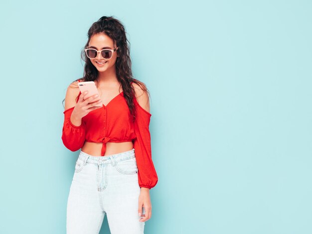 트렌디한 여름 빨간 상의와 청바지 옷을 입은 젊고 아름다운 웃는 여성 스튜디오의 파란색 벽 근처에서 포즈를 취하는 섹시한 평온한 여성 휴대폰 앱을 사용하여 스마트폰 화면을 보고 모델