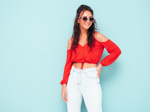 Young beautiful smiling female in trendy summer red top and jeans clothes Sexy carefree woman posing near blue wall in studio Positive brunette model having fun Cheerful and happy In sunglasses