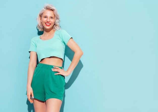 Young beautiful smiling female in trendy summer shorts and top clothes Sexy carefree woman posing near blue wall in studio Positive blond model having fun and going crazy Cheerful and happy