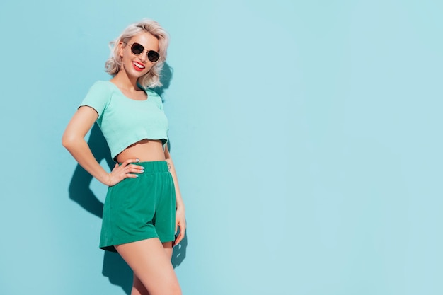 Young beautiful smiling female in trendy summer shorts and top clothes Sexy carefree woman posing near blue wall in studio Positive blond model having fun and going crazy Cheerful and happy