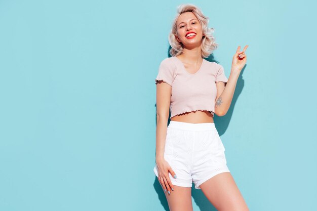 Young beautiful smiling female in trendy summer shorts and top clothes Sexy carefree woman posing near blue wall in studio Positive blond model having fun and going crazy Cheerful and happy