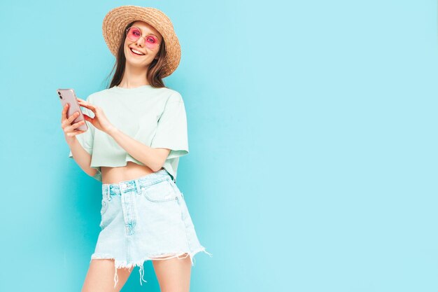 Young beautiful smiling female in trendy summer skirt Sexy woman with posing near yellow wall in studio Positive model having fun In sunglasses and hatLooking at smartphone Using apps
