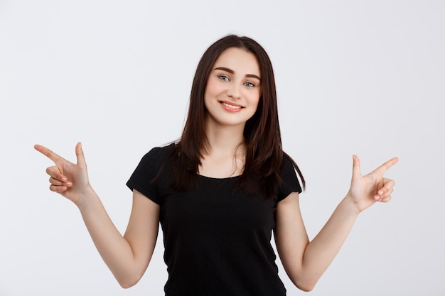 Young beautiful smiling girl in black t-shirt pointing fingers in opposite sides over white wall