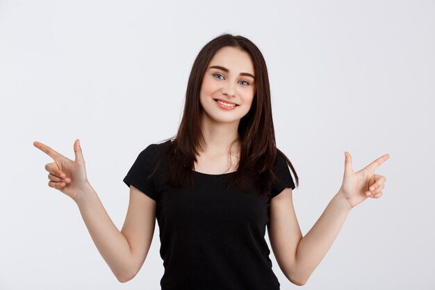 Молодая красивая улыбающаяся девушка в черной футболке, указывая пальцами в разные стороны над белой стеной