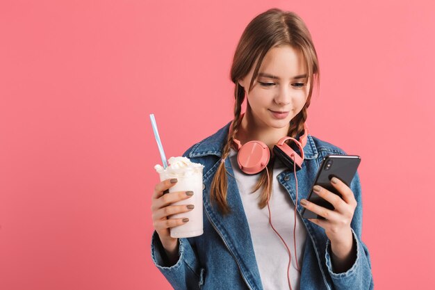 Young beautiful smiling girl with two braids in denim jacket with headphones holding milkshake in hand while dreamily using cellphone over pink background