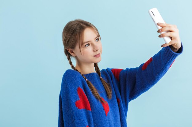 Young beautiful smiling girl with two braids in navy color oversize sweater dreamily taking photos on cellphone over blue background isolated
