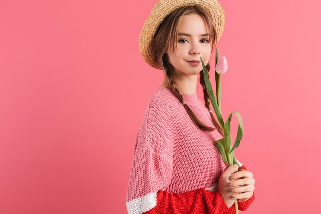 Young beautiful smiling girl with two braids in sweater and straw hat holding one tulip in hand dreamily looking in camera while spending time over pink background