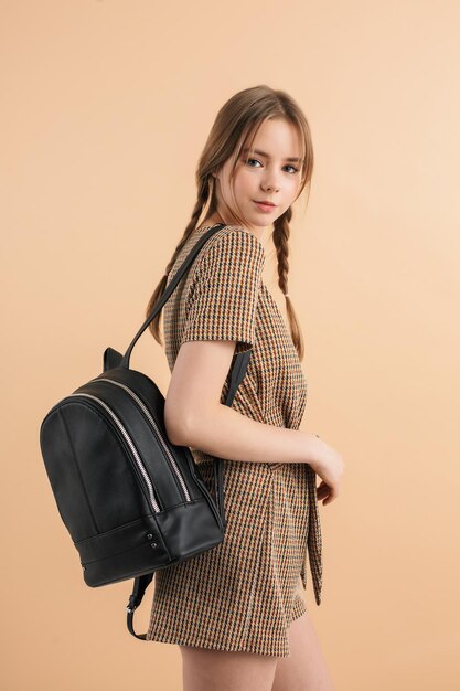 Young beautiful smiling girl with two braids in tweed jumpsuit holding black backpack on shoulder happily looking in camera while spending time over beige background
