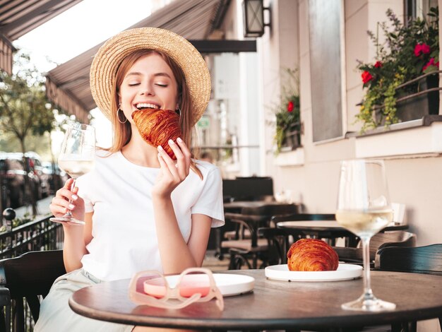 Young beautiful smiling hipster female in trendy summer clothesCarefree woman posing at veranda cafe in the streetPositive model drinking white wineEnjoying vacationEating croissant In hat
