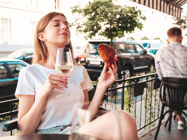 Young beautiful smiling hipster female in trendy summer clothesCarefree woman posing at veranda cafe in the streetPositive model drinking white wineEnjoying vacationEating croissant