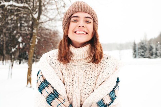 Молодая красивая улыбающаяся хипстерская женщина в модной теплой одежде и шарфе. Беззаботная женщина позирует на улице в парке. Позитивная чистая модель развлекается в снегу. Наслаждаясь зимними моментами