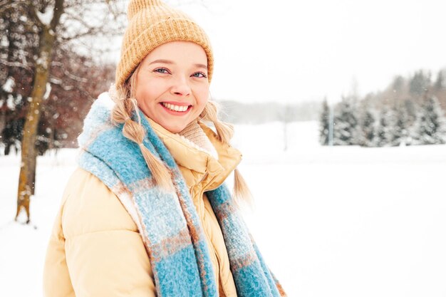 トレンディな暖かい服とスカーフで若い美しい笑顔のヒップスターの女性。公園の通りでポーズをとる屈託のない女性。雪の中で楽しむポジティブピュアモデル。冬のひとときを楽しむ