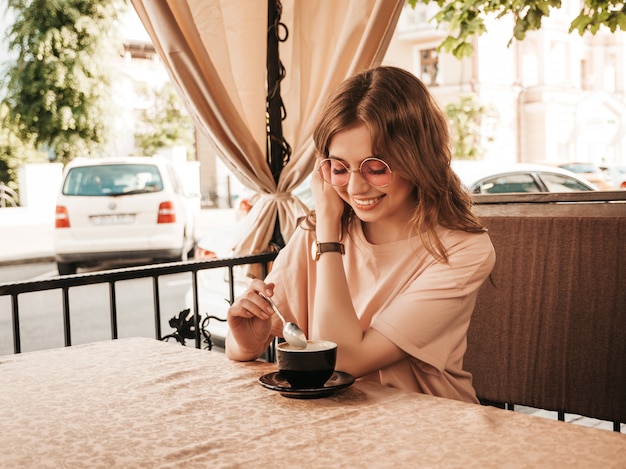 Young beautiful smiling hipster girl in trendy summer clothes.carefree woman sitting in veranda terrace cafe and drinking coffee.positive model having fun and dreams