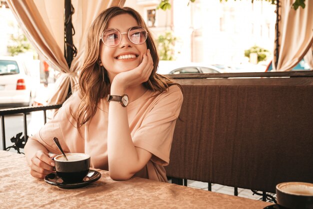 Молодая красивая улыбающаяся девочка битник в модной летней одежде. Беззаботная женщина, сидя в кафе на террасе веранды и пить кофе. Позитивная модель с удовольствием и мечты