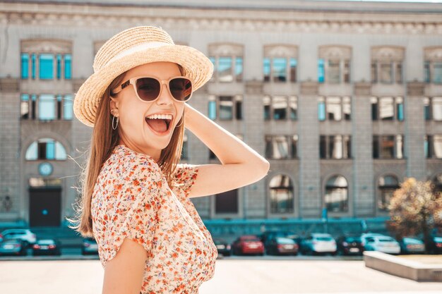 트렌디한 여름 옷을 입은 젊고 아름다운 미소 힙스터 여성 해질녘 거리 배경에서 포즈를 취하는 섹시하고 평온한 여성 야외 긍정적인 모델 선글라스와 모자에 쾌활하고 행복합니다.
