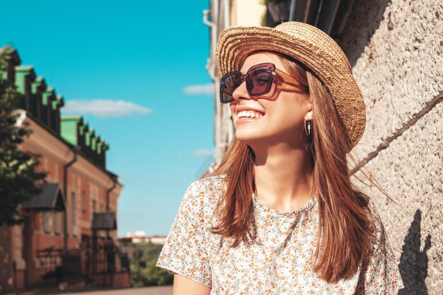 トレンディな夏の服を着た若い美しい笑顔の流行に敏感な女性日没時に通りの背景でポーズをとるセクシーな屈託のない女性屋外のポジティブモデルサングラスと帽子で陽気で幸せ