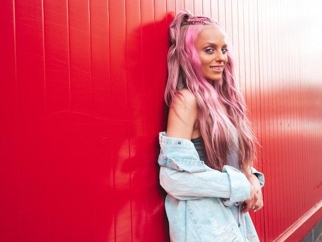 Young beautiful smiling hipster woman in trendy summer jeans shorts with pink hairstyle and makeup. Sexy carefree model posing in the street near red wall. Positive model outdoors