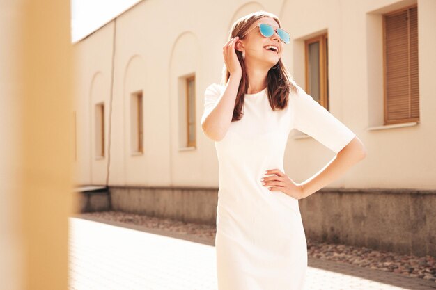 Молодая красивая улыбающаяся хипстерша в модном летнем белом платье Сексуальная беззаботная женщина позирует на фоне улицы на закате Позитивная модель на улице Веселая и счастливая в солнечных очках