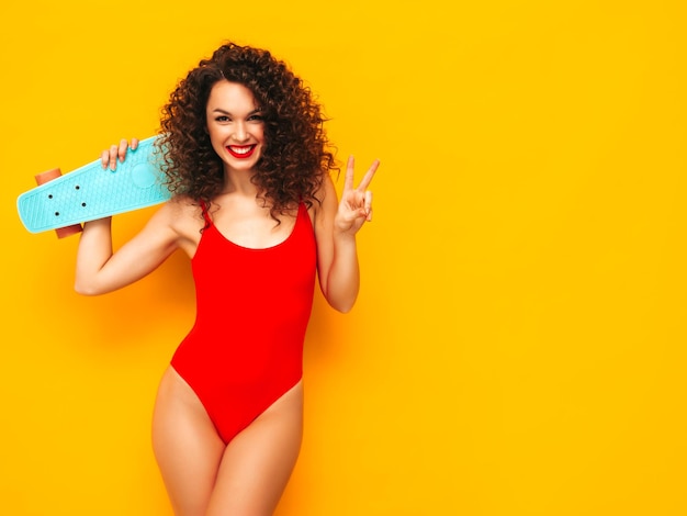 Young beautiful smiling woman posing near yellow wall in studioSexy model in red swimwear bathing suitPositive female with curls hairstyle Holding penny skateboardHappy and cheerful