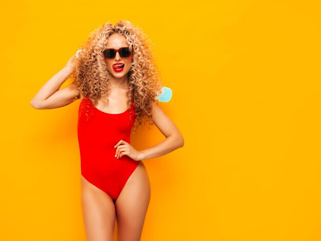 Young beautiful smiling woman posing near yellow wall in studioSexy model in red swimwear bathing suitPositive female with curls hairstyle Holding penny skateboardHappy and cheerful