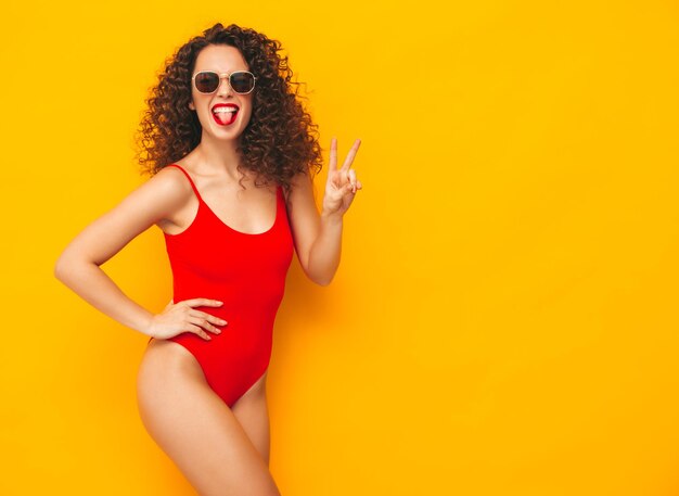 Молодая красивая улыбающаяся женщина позирует возле желтой стены в студииСексуальная модель в красном купальном костюмеПозитивная женщина с кудрявой прическойСчастливая и веселая В солнцезащитных очках Показывая знак мира