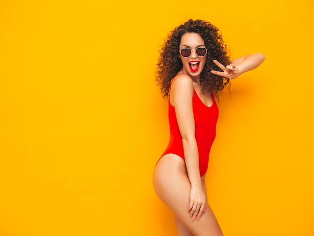 Молодая красивая улыбающаяся женщина позирует возле желтой стены в студииСексуальная модель в красном купальном костюмеПозитивная женщина с кудрявой прическойСчастливая и веселая В солнцезащитных очках