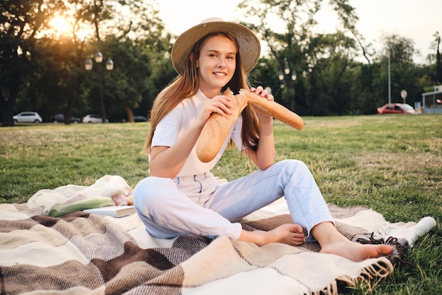 Young beautiful smiling woman in straw hat sitting on plaid with baguette bread while dreamily looking in camera on picnic in city park