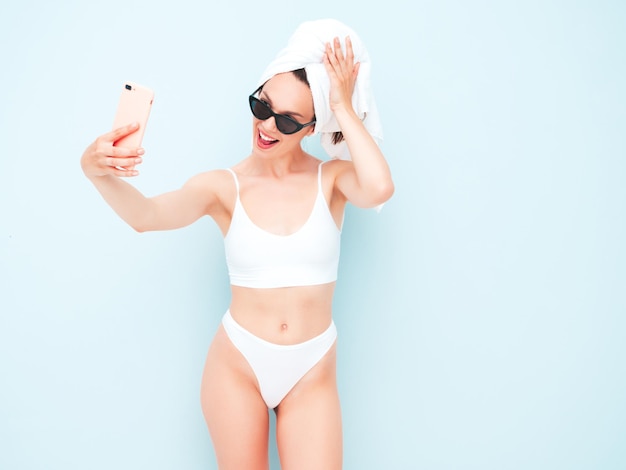 Young beautiful smiling woman in white lingerie. Sexy carefree model in underwear and towel on head posing pink wall in studio. Positive and happy female enjoying morning. Taking selfie photos