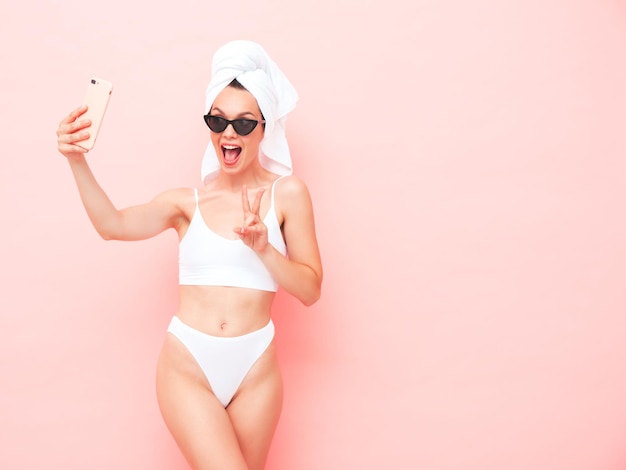 Young beautiful smiling woman in white lingerie. Sexy carefree model in underwear and towel on head posing pink wall in studio. Positive and happy female enjoying morning. Taking selfie photos