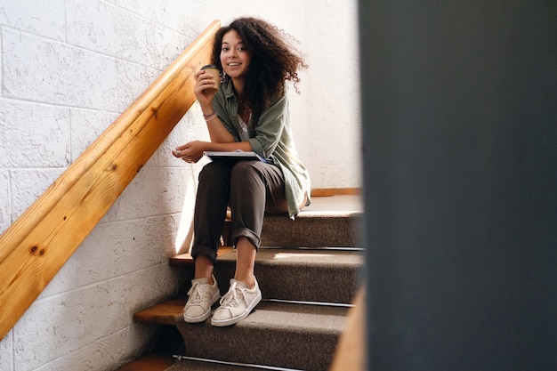 Young beautiful smiling woman with dark curly hair sitting on stairs in university with textbooks on knees and cup of coffee to go in hand happily looking in cameraxA