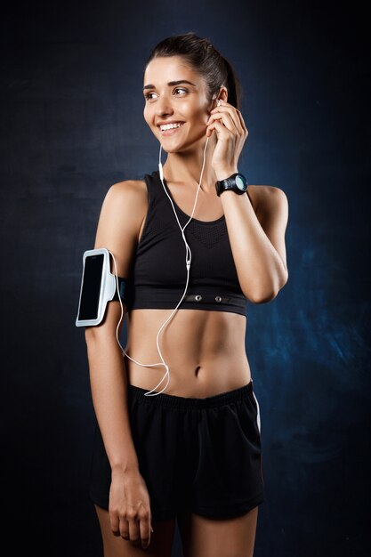 Young beautiful sportive girl listening music over dark wall.