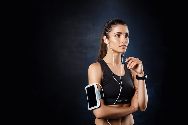 Young beautiful sportive girl listening music over dark wall.