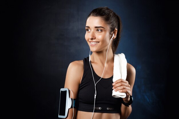 Young beautiful sportive girl listening music over dark wall.