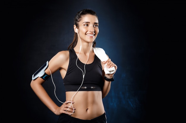 Young beautiful sportive girl listening music over dark wall.