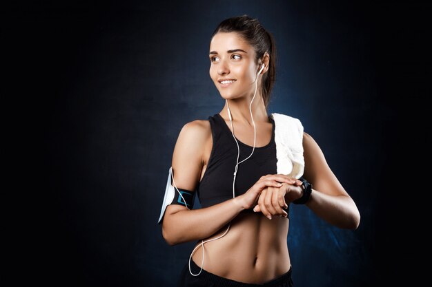 Young beautiful sportive girl listening music over dark wall.