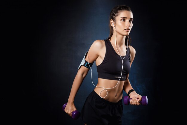 Young beautiful sportive girl training with dumbbells over dark wall.