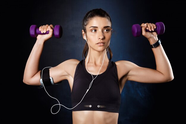 Young beautiful sportive girl training with dumbbells over dark wall.