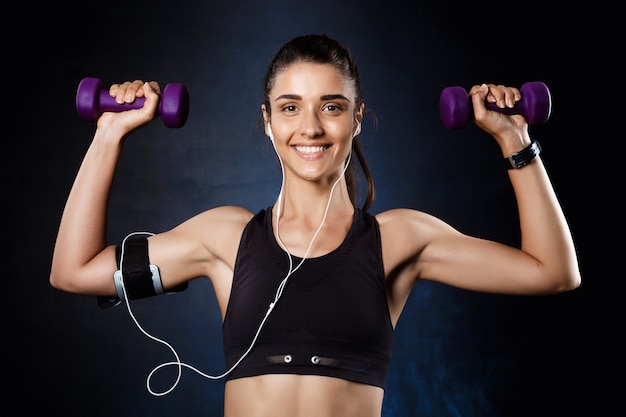 Young beautiful sportive girl training with dumbbells over dark wall.