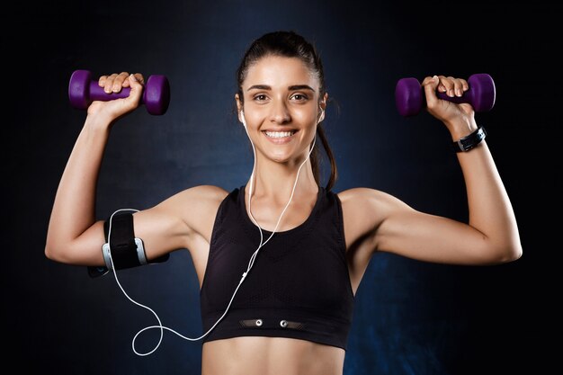 Young beautiful sportive girl training with dumbbells over dark wall.