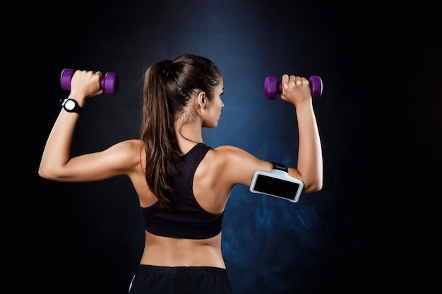 Young beautiful sportive girl training with dumbbells over dark wall.