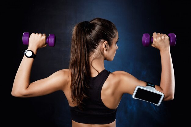 Young beautiful sportive girl training with dumbbells over dark wall.