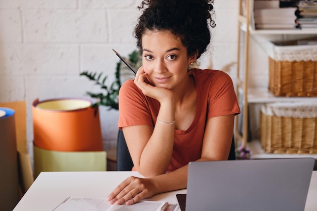 Young beautiful student girl with dark curly hair sitting at the table with laptop leaning on hand happily looking in camera studying at modern cozy home