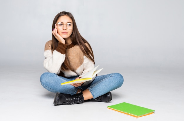 Young beautiful student sitting with book, reading, learning. Isolated on white wall