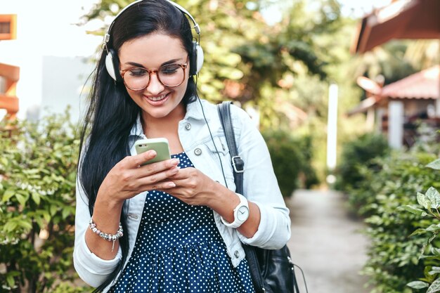 Young beautiful stylish woman using smartphone, headphones, eye glasses, summer, vintage denim outfit, smiling, happy, positive