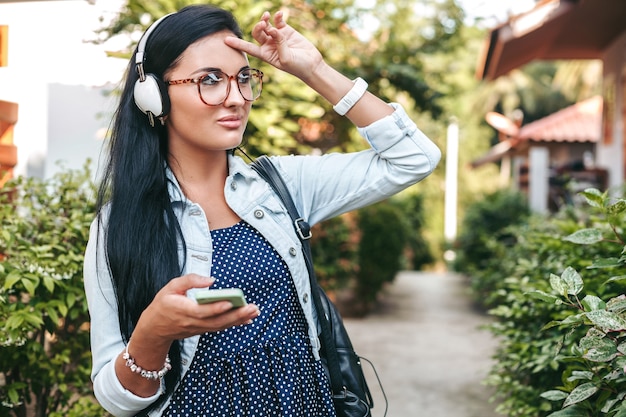 Foto gratuita giovane bella donna elegante che utilizza smartphone, cuffie, occhiali da vista, estate, vestito di denim vintage, sorridente, felice, positivo