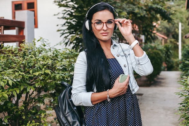 Young beautiful stylish woman using smartphone, headphones, eye glasses, summer, vintage denim outfit, smiling, happy, positive