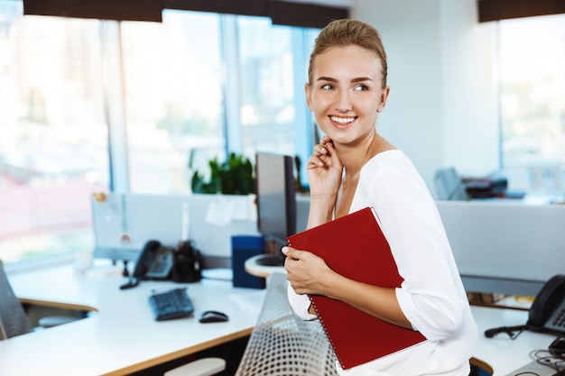 Young beautiful successful businesswoman smiling, posing, holding folder, over office