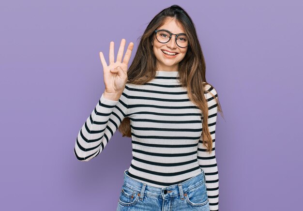 Молодая красивая девушка-подросток в повседневной одежде и очках показывает и указывает пальцами номер четыре, улыбаясь уверенно и счастливо