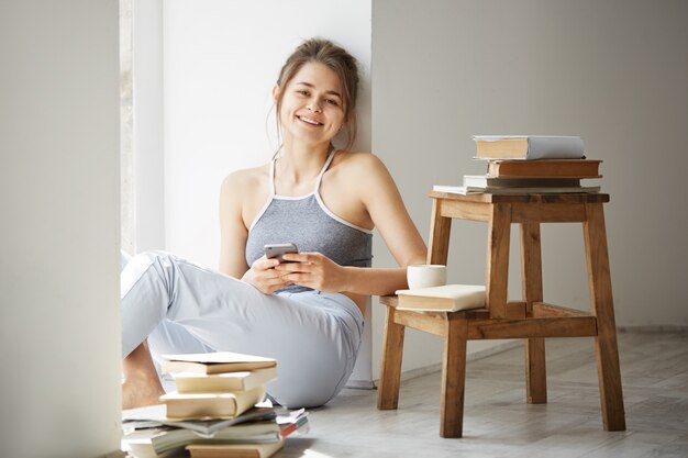 Young beautiful teenage woman surfing internet at phone smiling sitting on floor among old books near window over white wall.